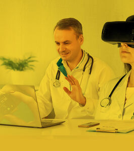 Médico sentado à mesa com portátil à sua frente acena para o portátil, demonstrando uso de tecnologia em teleconsulta, e médica com óculos de realidade virtual ao lado, ilustração sobreposta ao portátil de uma relógio digital com dados de saúde 